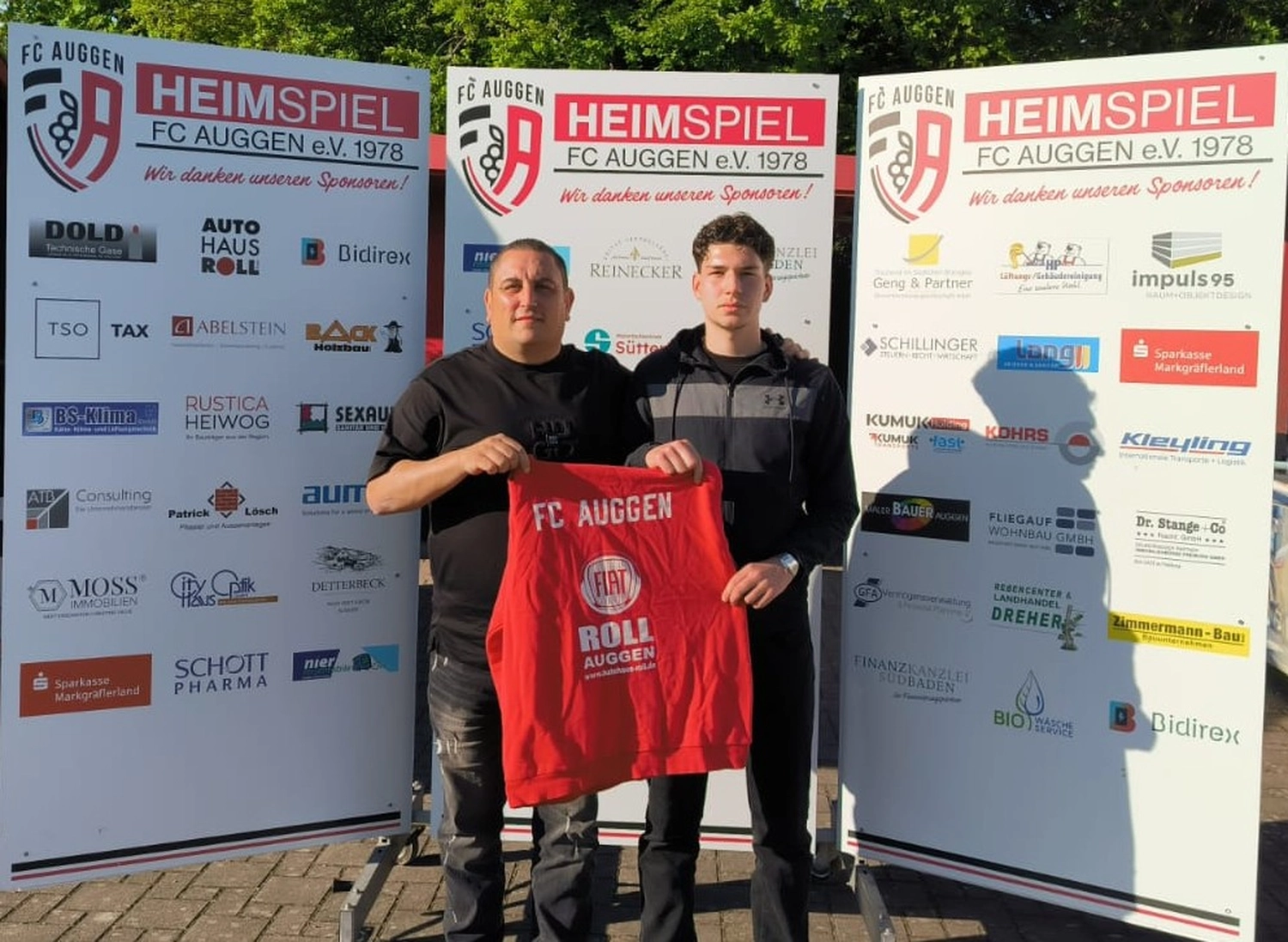 FC Auggen e.V. – Fußballtrainer – Dieses Bild zeigt ein rotes Sweatshirt mit den Logos von FC Auggen, Fiat und Autohaus Roll, das von zwei Personen gehalten wird.